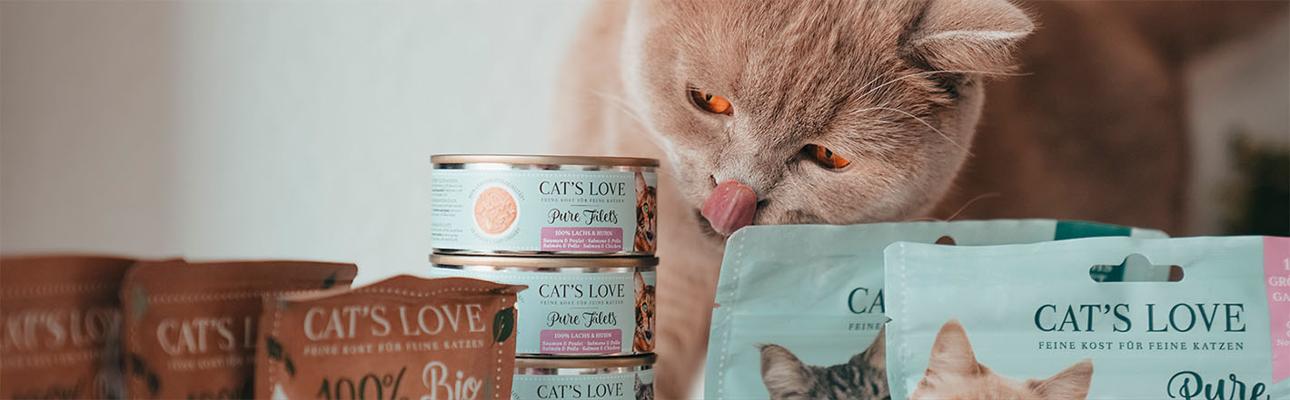 A cat sitting behind different CAT'S LOVE products and licking its mouth
